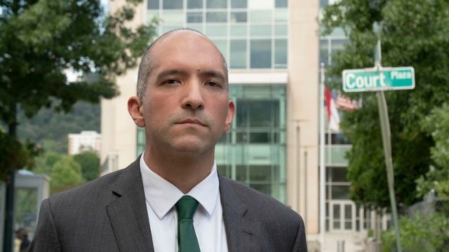 Public defender Ehsan Akhavi, a light skinned young bald man in a business suit outside a building with a road sign behind him that reads "Court Plaza"
